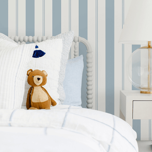 A blue and white-striped wallpaper in a child's room with white bedding and stuffed toy