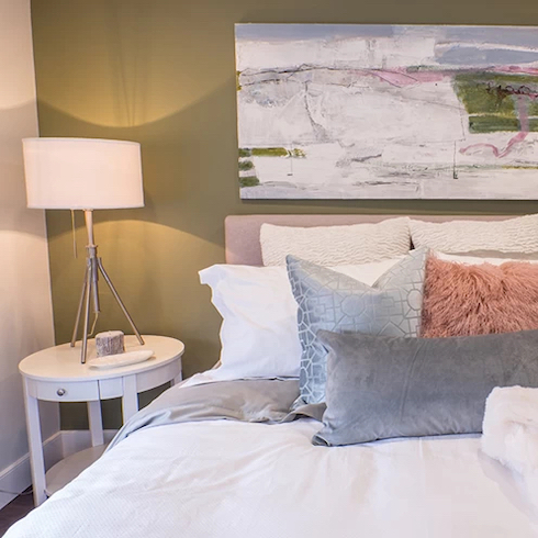 Beautiful bedroom with a sage green feature wall and large piece of art, a pink bed with white and grey bedding and a small round bedside table with a white lamp as featured on HGTV Canada.