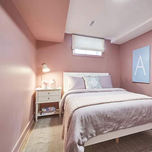 Basement bedroom from Extreme Makeover: Home Edition on HGTV Canada is painted in all over petal pink with a double bed with pink and white bedding and a side table and area rug