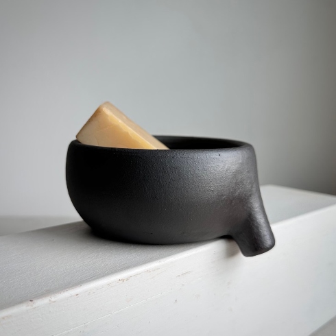 A black clay soap dish with a built-in strainer sitting on the edge of a sink.