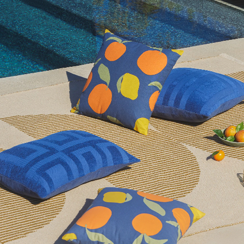 Blue and citrus-patterned outdoor pillows lay near a pool on a sunny summer day