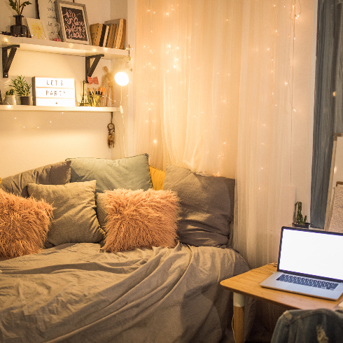 A dorm room lit up with string lights with plenty of accent pillows and an open shelf