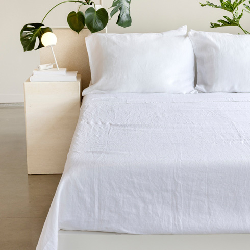 White sheets on a bed with a tall green plant and side table next to it.