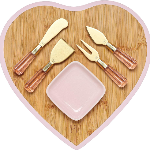 A wooden pink-trimmed heart-shaped charcuterie board with gold and pink accessories