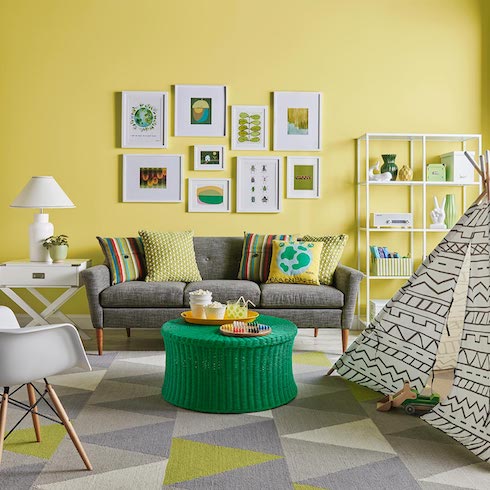 Funky living room with a white plastic molded chair, bright green rattan coffee table, grey couch, white and black play tent, white metal shelves, white table lamp on a side table and a gallery of framed art on the wall painted in Sherwin-Williams SW 0073 Chartreuse