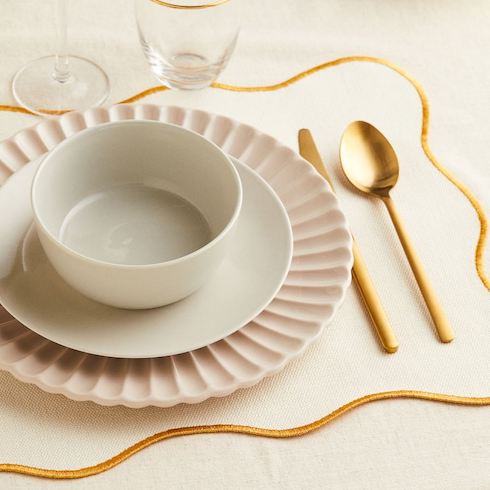 Cream and gold-colored placemat made from woven cotton fabric with scalloped edges with contrasting stitching as part of a pretty Thanksgiving table setting with scalloped charger, white plate and bowl and golden flatware from H&M Home