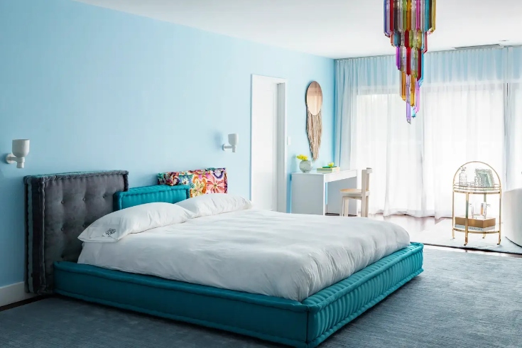 A bedroom with pale aqua walls, a large window, and a bed with turquoise-coloured velvet frame
