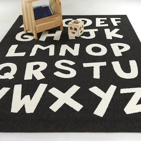 Mod-Tod Beta Kids Alphabet Indoor/Outdoor Area Rug in Charcoal available at Bed Bath & Beyond shown on the floor with a child’s shape sorter toy and a wooded book box as featured in HGTV Canada's Best Nursery Rugs Under $200 Your Little One This Year