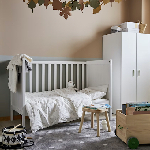 Grey rug with white dots from Ikea in a baby’s nursery decorated with a fall leaf themed bunting, white toddler bed, white wardrobe, a drum, a wooden stool and a wheeled wooden book box as featured in HGTV Canada's Best Nursery Rugs Under $200 Your Little One This Year