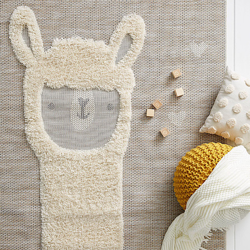 Sweet faced llama rug from Simons on a white wooden floor with a white blanket, yellow pouf, white pillow and wooden blocks laying on top of it as featured in HGTV Canada's Best Nursery Rugs Under $200 Your Little One This Year