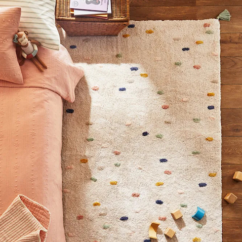 Cotton rug with multicolored polka dots and a non-slip base from Zara shown in a child’s nursery with wooden blocks, a floor bed with pink sheets and wicker bedside table as featured in HGTV Canada's Best Nursery Rugs Under $200 Your Little One This Year