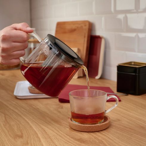 Glass teapot pouring tea into cup on wood counter.