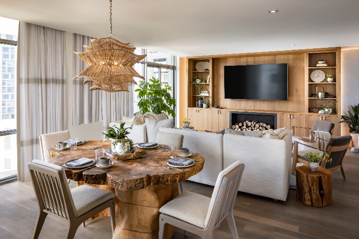 A combination living and dining area in a hotel suite with rattan pendant light, live edge wooden table, chairs, sofa and a wall-mounted TV on light coloured wood panelling.