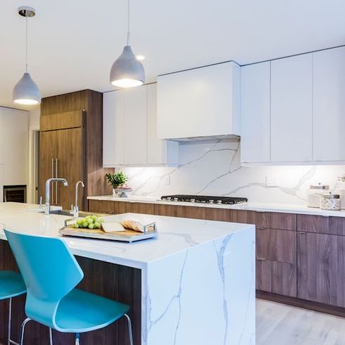 A chic kitchen designed by Hilary Farr melding patterned wood, streaky marble, concrete pendant lamps, matte white cabinetry and two teal bar stools