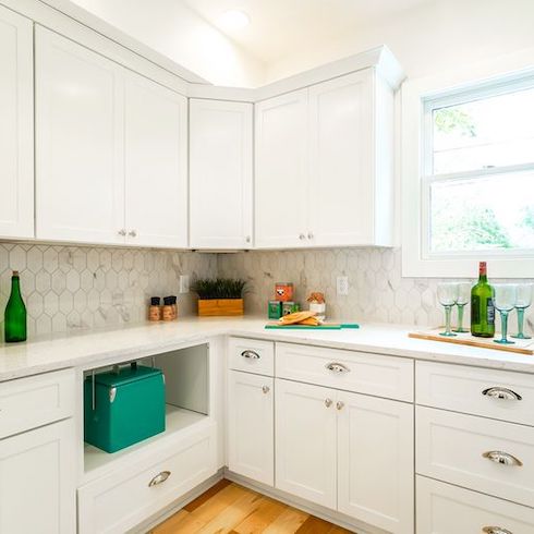 Roomy, pristine prep kitchen renovation from Masters of Flip stars Dave and Kortney Wilson for HGTV Canada featuring white floor to ceiling cabinets, a perfectly placed window, and a white marble topped counter with green bottles, a cutting board, tea tins, wine glasses, spices and a potted plant