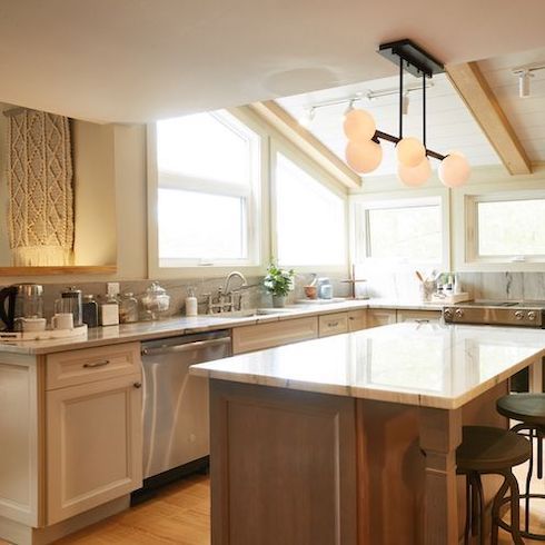 A bright white kitchen featuring new, energy-efficient windows, a multi globe pendant light over the kitchen island, white shaker style cabinets, white shiplat and exposed beam ceiling, and white marble countertops designed by Scott McGillivray for HGTV Canada’s Scott’s Vacation House Rules kitchen