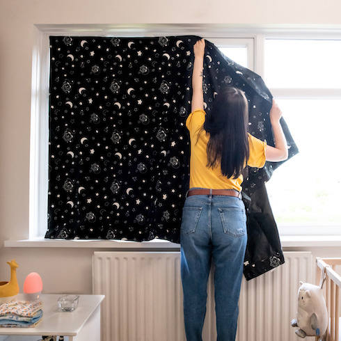 Woman in a yellow shirt hangs a starry night printed Tommee Tippee Portable Blackout Blind up on a window in a small nursery room with a wooden crib, a white radiator, and side table with baby muslins cloths, a pink night light and a small giraffe basket