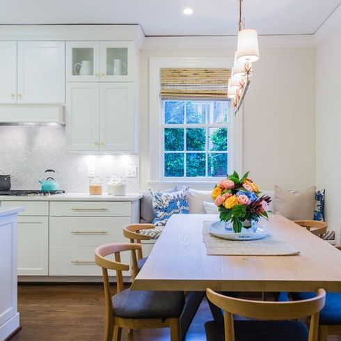A comfy, family-forward kitchen and dining area designed by Hilary Far featuring a teak dining set, piles of pillows, rattan blinds, white cabinetry and fresh bouquet of flowers on the table