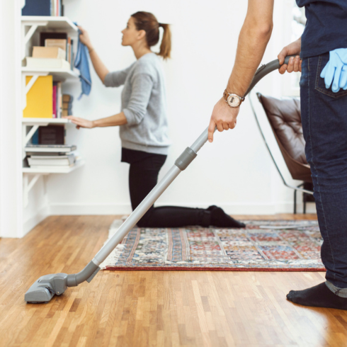 couple cleaning apartment; woman dusting bookshelf while man cleans hardwood floor with dry mop