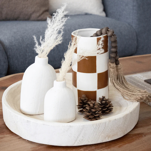 Gift idea - Wyrth ceramic Checkerboard Vase with a wooden beaded string draped on its top edge sits on a white wooded tray with two small ceramic petal vases with white grasses and three pinecones on top of a wooden coffee table in front of a grey couch