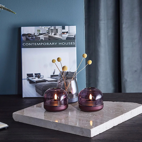Gift idea - Two Kooduu Flameless Candles Glow 8, made of a coloured purple glass hurricane on aluminum base, and a smokey gree Koodoo Flameless Candle Glow 10 with dried flowers in it, sit on a piece of grey marble on top of a dark wood table along with a large coffee table book titled “Contemporary Houses”