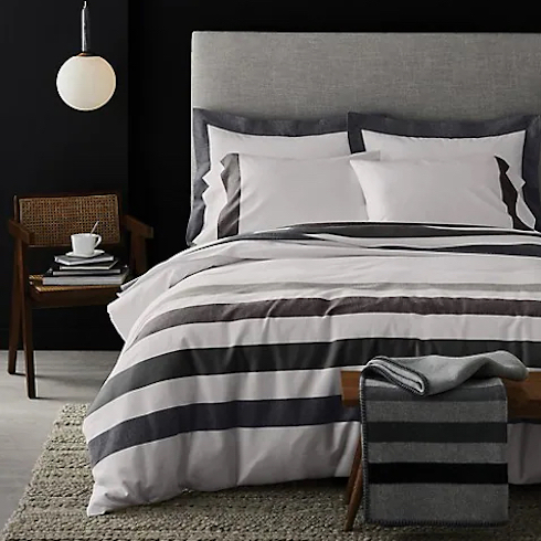 HBC Signature Sterling Stripe Flannel Duvet Cover Set on a large grey upholstered bed in a chic black bedroom with grey floors and a knitted rug, a cane chair with a stack of books and white cup and saucer, and a white pendant lamp hanging from the ceiling