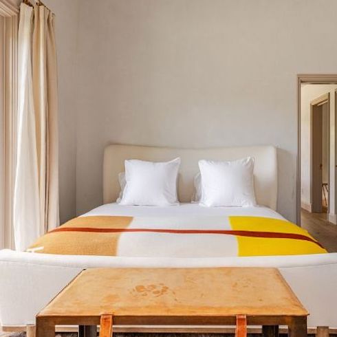 Celebrity homes - A luxurious white bedroom in Gwyneth Paltrow’s Montecito Guesthouse with cream coloured drapes, a white queen size bed, bold yellow blanket and a leather topped table at the foot of the bed