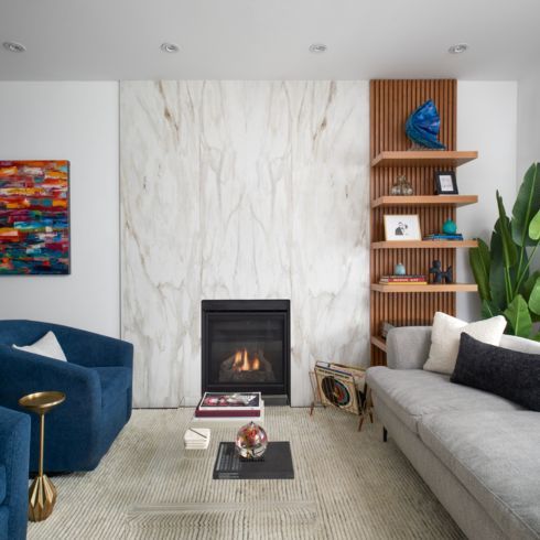Stunning modern living room with a black insert fireplace surrounded by floor to ceiling porcelain tiles, cream carpet, blue armchairs, grey couch, colourful wall art, wooden display shelf unit, and a large potted palm