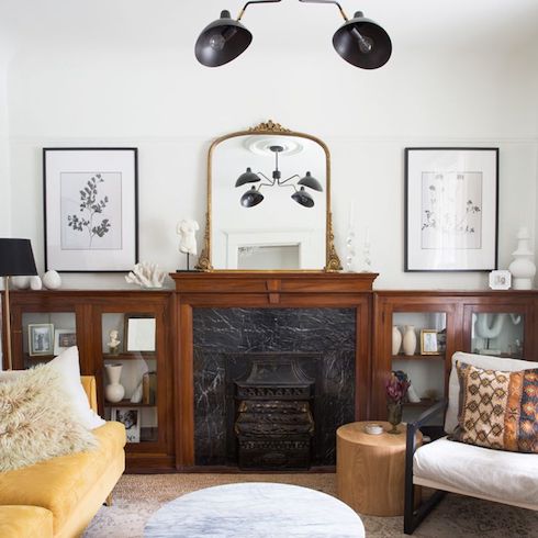 Historic fireplace mantel and side cases adorned with vintage pieces, art, books and gold French mirror in a living room designed by Lauren Miller featuring a yellow couch, botanical prints on the wall, a stump stool, a linen armchair, and a round marble table