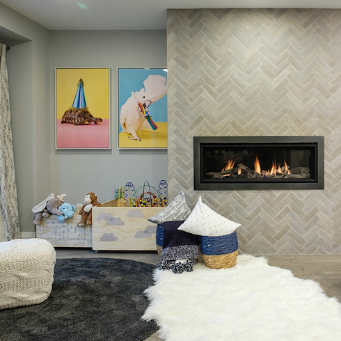A chic modern basement play area features a long gas fireplace, a herringbone tile walls, two full toy boxes, a knitted pouf, two baskets with pillows, a grey round rug, a white faux fur rug, and grey walls with two bright paintings