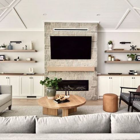 Electric fireplace with a thick wooden mantel and TV mounted above it in a charming living space with white side cabinets and two sets of floating shelves on either side, a large couch, a round coffee table with books and a botted plant, two black framed armchairs, a leather pouf, and a cream coloured rug