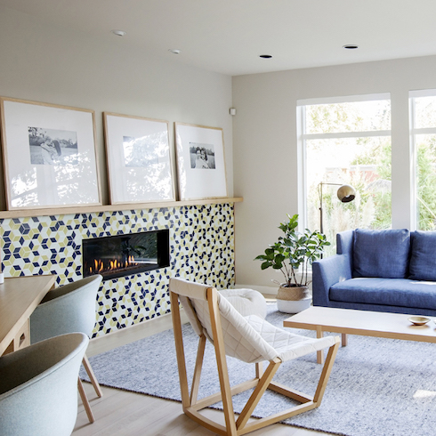 Modern living room with colourful blue sofa, white quilted chair, rectangular wooden coffee table, a large potted plant, two big windows, blue rug and tiled fireplace mantel with three big framed photos