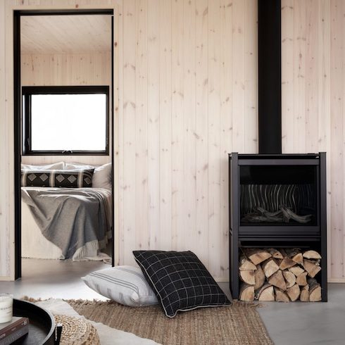 Cozy sitting area with two large floor cushions, a jute rug, a black standalone Scandinavian woodstove against a pine panelled wall looking through into a bedroom with a made bed and a black framed window