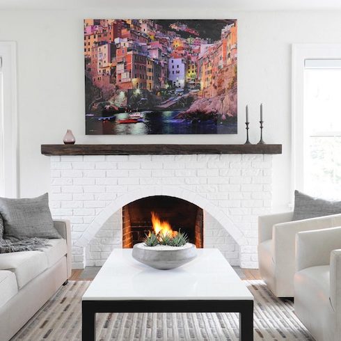 Minimal white living room with colourful painting above mantel, an arch white brick fireplace surround, a white couch, two white armchairs, a white mosaic rug, a white and black rectangular coffee table with a succulent bowl on top, and a fire in the open wood burning fireplace