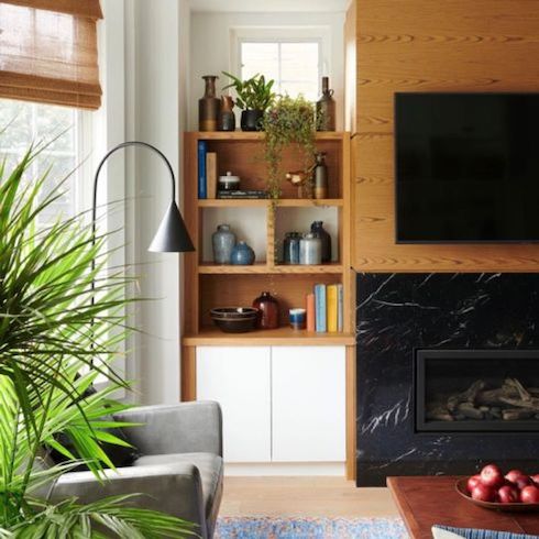Gas fireplace with a black marble surround flanked by walnut wood shelves to the side filled with books, plants and vases, and above with a mounted TV, in a living room with a grey armchair, a large potted palm, and a wooden coffee table with a bowl of red apples