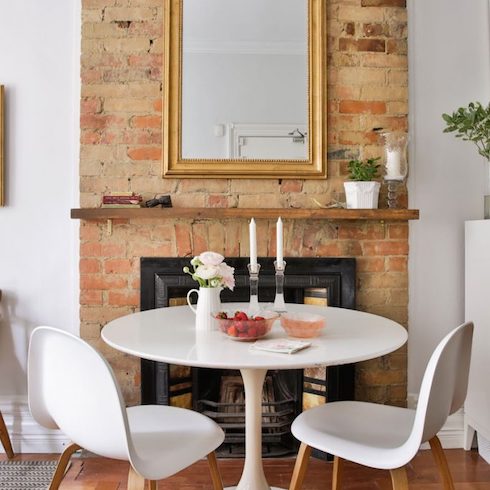 Fireplace with original bricks and inlay, a wooden mantel, a gold framed mirror on the wall, a white round breakfast table and two white chairs feature in a home redesign by Lauren Miller