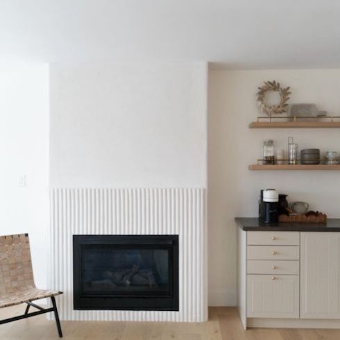 White reeded fireplace in modern room with a woven occasional chair and side counter with a tea and coffee station and two floating wooded shelves above