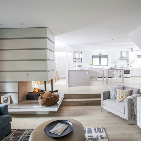 A living room with a three-way fireplace in a contemporary and lavish designed by Jillian Harris with white oak flooring, a big grey armchair, two steps up to a large modern, white kitchen and dining area