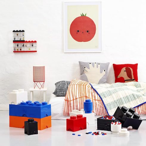 Styled kids bedroom with Lego brick storage containers on the floor, a single bed with bright bedding, and a tomato print on the white wall behind