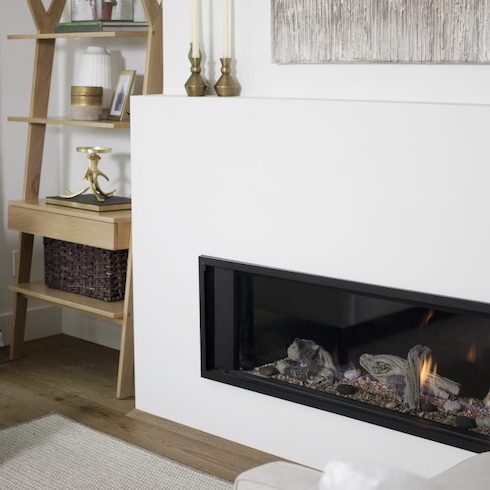 Indoor linear fireplace surrounded by white stone with a shallow mantle holding two brass candlestick holders, oak flooring, a white rug, and a corner ladder shelf holding books and art designed by Janis Nicolay