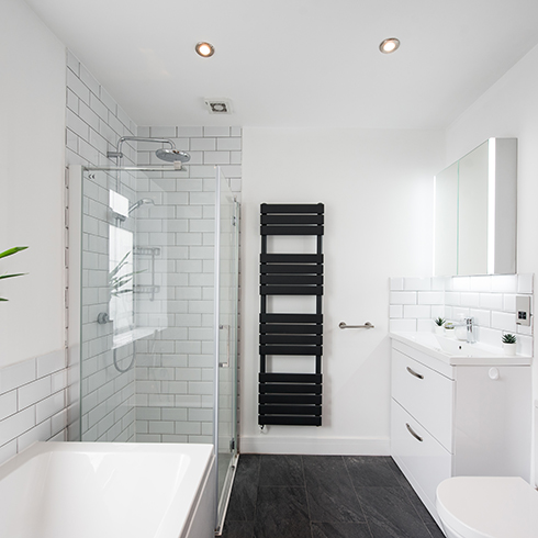 A general interior view of a white half rectangular tiled and painted bathroom, bathtub, glass shower cubicle, chrome taps and shower head, window and potted dragon tree plant, sink mounted to a vanity unit, mirror medicine cabinet and black towel warmer radiator and a picture frame with the words saying 