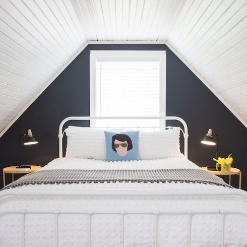 A guest black and white bedroom with a window, white wood panelled walls and ceiling, a white metal bedframe with white bedding, two yellow side tables, two black wall scones, and an Elvis Presley throw pillow