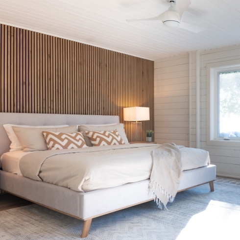 Chic luxurious bedroom with a slat black and wood feature wall, white panelled walls and ceiling, a white ceiling fan, a large grey upholstered bed with tan bedding and two patterned throw pillows, a side table with a glass lamp and small potted plant
