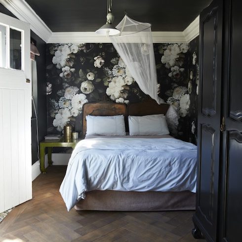 Moody black floral wallpaper in bedroom with dark chevron parquet flooring, a large black armoire, a white mosquito net, a green metal side table, a wood and linen bedframe with white sheets and pillows, a metal pendant lamp, a black ceiling with white molding, and a white exterior door