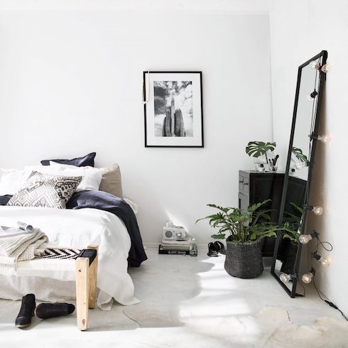 Black and white bedroom in a chic loft with a low bed with white and black linens, a wooden bench with a pair of black boots underneath it, a concrete floor with a white skin rug, a potted plant in a black basket, a black framed mirror with a set of string lights leaning against the white walls, a small black dresser with a small potted palm, a pile of books on the floor and a framed black and white photo on the ground