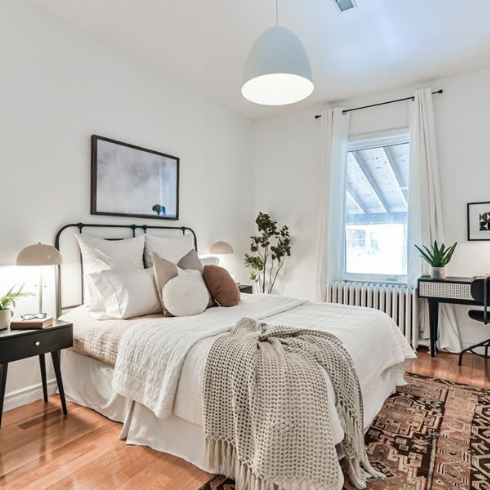 Beautiful staged bedroom in a Toronto house for sale near High Park featuring newly refinished wood floors, white walls, black furniture, black framed artwork, black metal framed bed with white and cream bedding, and a large black and rose coloured Persian rug