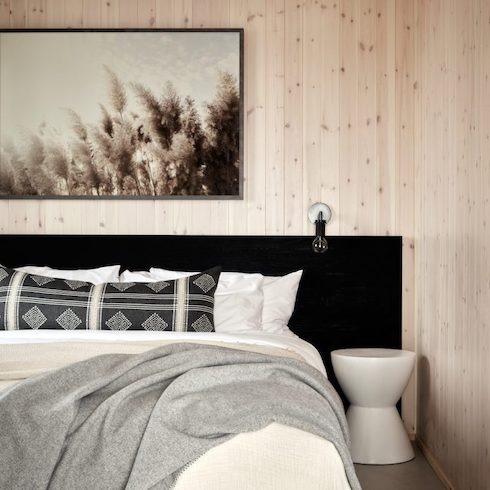Guest bedroom at Wander the Resort featuring an extra wide black headboard, large grass artwork, light wood wall panelling, bed with white sheets and black pillows, and a drum bedside table