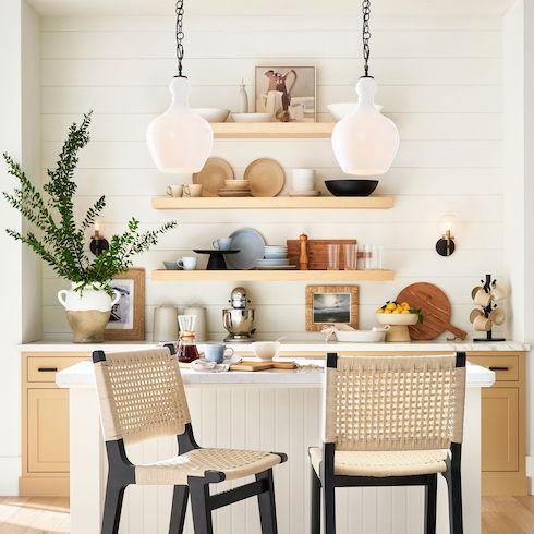 Two Flynn Milk Glass Pendants by Pottery Barn, featured in HGTV Canada’s kitchen lights gallery hang in a small breakfast nook kitchen with wooden lower cabinets with black hardware, white marble countertop, three floating wooden shelves filled with bowls and plates, a small centre island with white panelling and two wicker and black bar stools