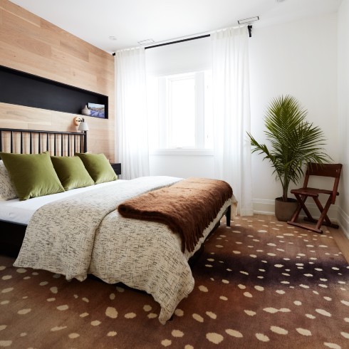 Chic bedroom with a large brown and cream spotted rug, large black framed bed with a brown throw, white sheets and three green pillows, a wood feature wall with a black insert shelf, a large potted palm, a wooden occasional chair, light white drapes and a black curtain rod