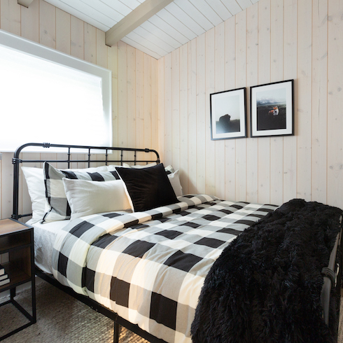A black and white bedroom in a Muskoka cabin featuring black and white buffalo check sheets, a black faux fur throw, white oak panelled walls, a black metal bedframe, a window, a wood and metal bedside table, and two black and white framed photos on the wall
